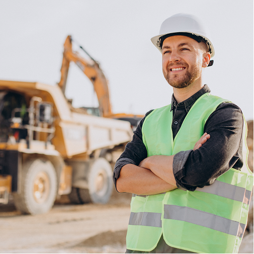 A construction worker wearing a white hard hat and reflective safety vest stands with arms crossed, smiling at a construction site with heavy machinery in the background.