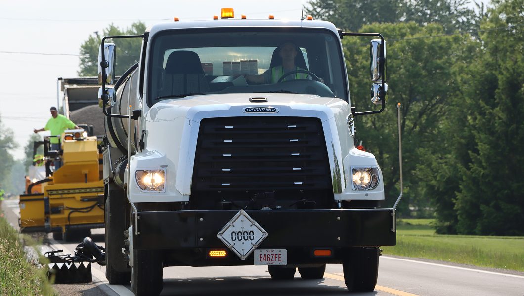 A white utility truck with hazard placards drives on a rural road, followed by road construction equipment and workers in high-visibility clothing. Trees and grass line the roadside.