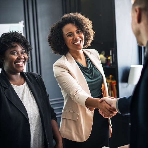 Two women in business attire smiling, with one of them shaking hands with a man in an office setting.