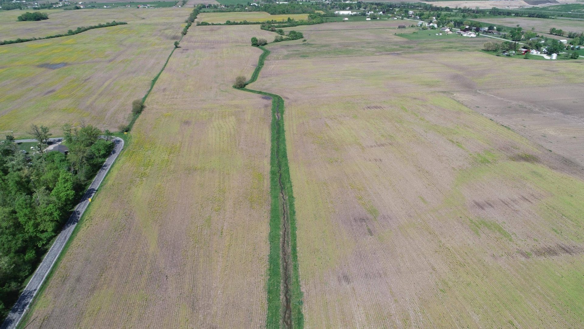 Aerial view of large, flat farm fields with a narrow green strip running through the center. Roads and trees line the edges, and scattered houses are visible in the distance.