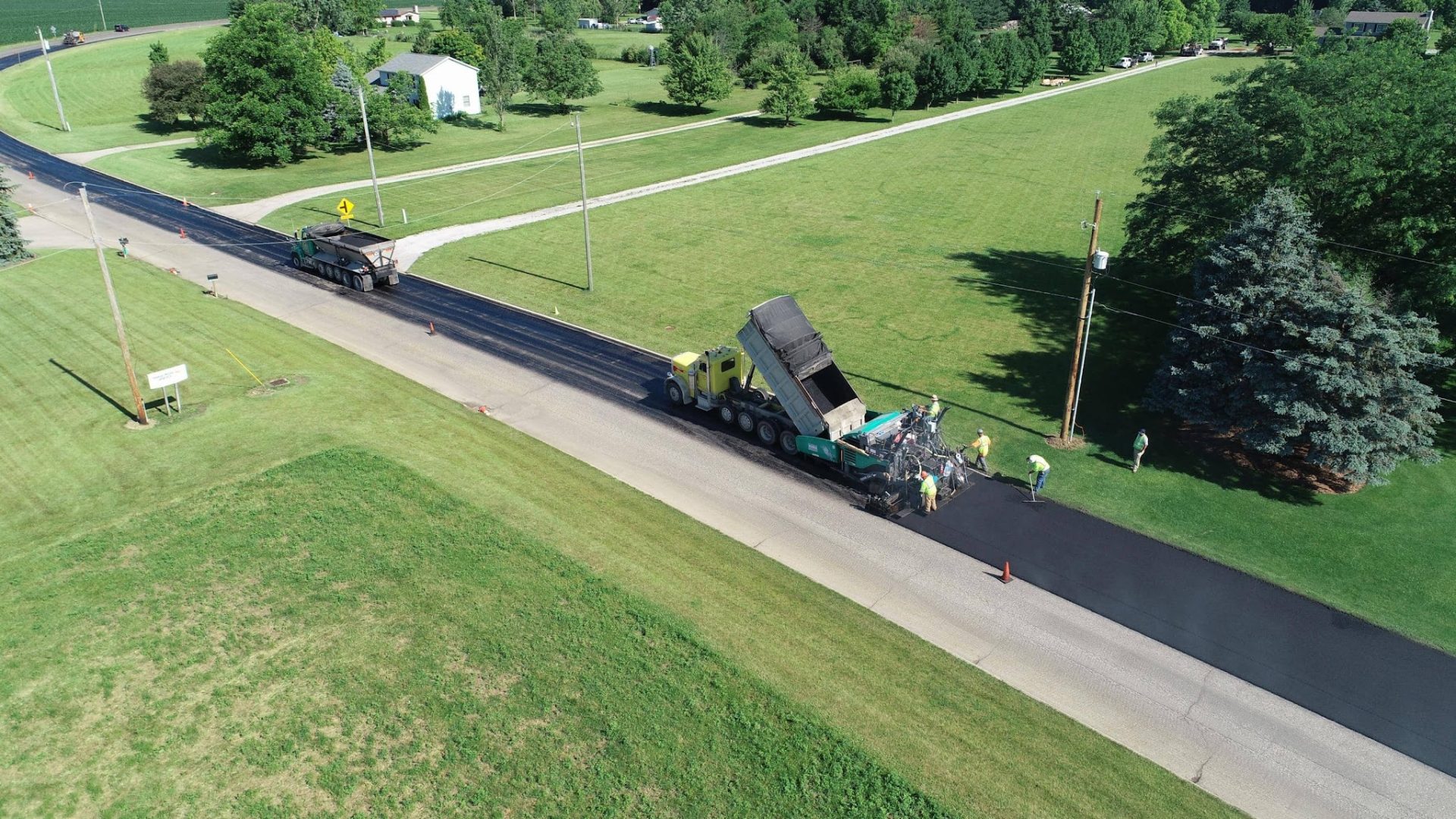 Aerial view of a road being paved by construction workers and heavy machinery, with green lawns, trees, and a white house in the background on a sunny day.