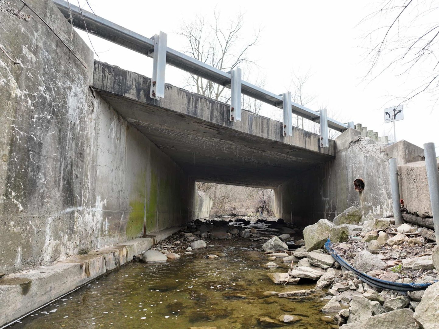 A concrete bridge crosses over a shallow creek with scattered rocks and stones. The bridge has metal guardrails and some moss or discoloration on its side. Leafless trees are visible in the background. A concrete bridge crosses over a shallow creek with scattered rocks and stones. The bridge has metal guardrails and some moss or discoloration on its side. Leafless trees are visible in the background.