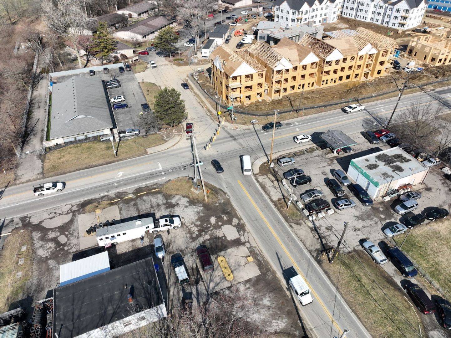 Aerial view of an intersection with cars, parking lots, and buildings, including a construction site with wooden-framed townhouses and several completed white buildings nearby. Trees and streets surround the area.