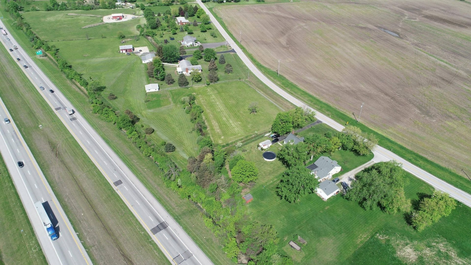 Aerial view of a rural area with houses, green lawns, trees, and fields. A highway with cars runs on the left, while smaller roads and houses are on the right, surrounded by open farmland.