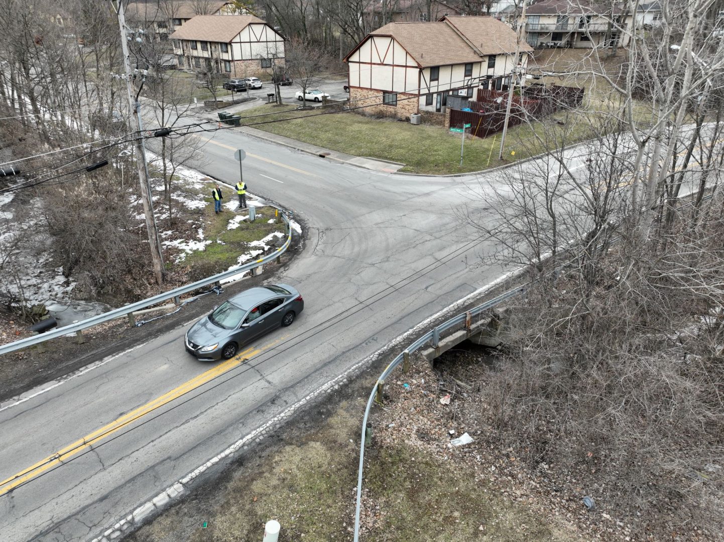 Aerial view of a car driving on a curved road intersection surrounded by leafless trees and houses; three workers in yellow vests stand near cables and utility boxes at the corner. Aerial view of a car driving on a curved road intersection surrounded by leafless trees and houses; three workers in yellow vests stand near cables and utility boxes at the corner.