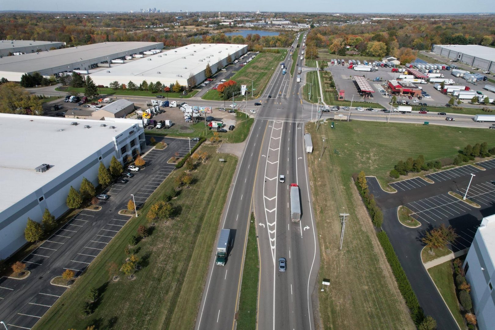 Aerial view of a wide road with trucks and cars, surrounded by large industrial buildings, parking lots, and green spaces, with a city skyline visible in the distant background.