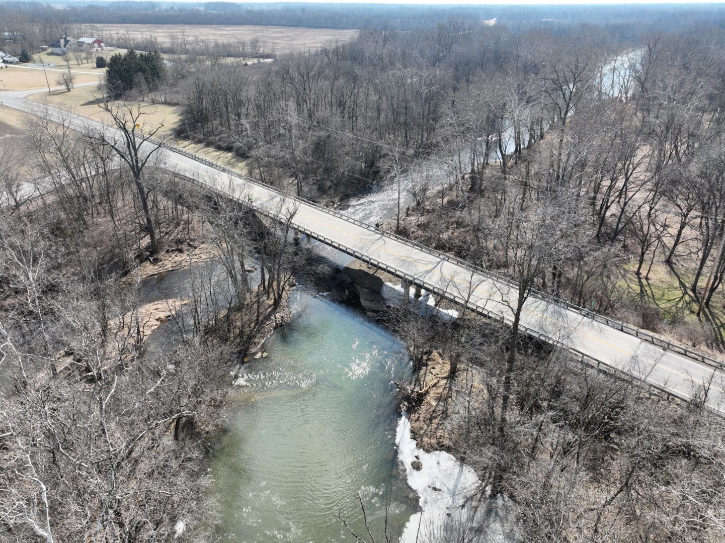 Aerial view of a rural road bridge crossing over a winding river surrounded by leafless trees and open fields on a clear day.