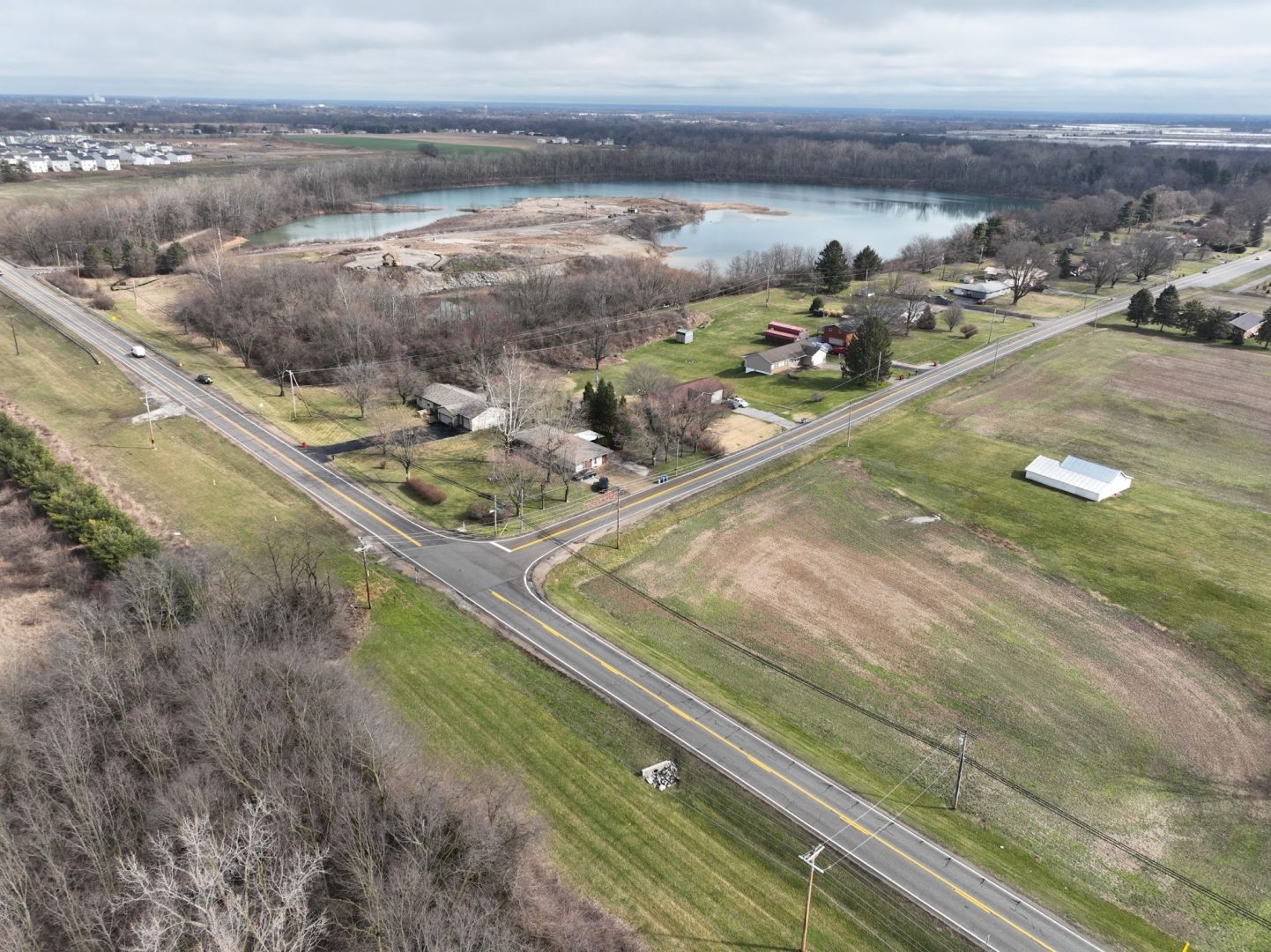 Aerial view of a rural intersection surrounded by fields, homes, and a large pond with trees. Roads cross in the foreground, with scattered houses and open land under a cloudy sky.