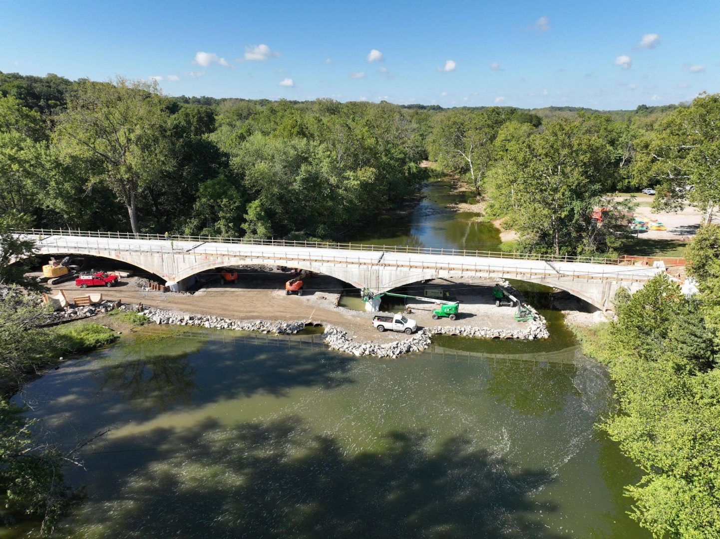 A concrete bridge spans a river surrounded by dense green trees; construction vehicles and equipment are visible on the riverbank, with rocks lining the shore under a blue sky with scattered clouds. A concrete bridge spans a river surrounded by dense green trees; construction vehicles and equipment are visible on the riverbank, with rocks lining the shore under a blue sky with scattered clouds.