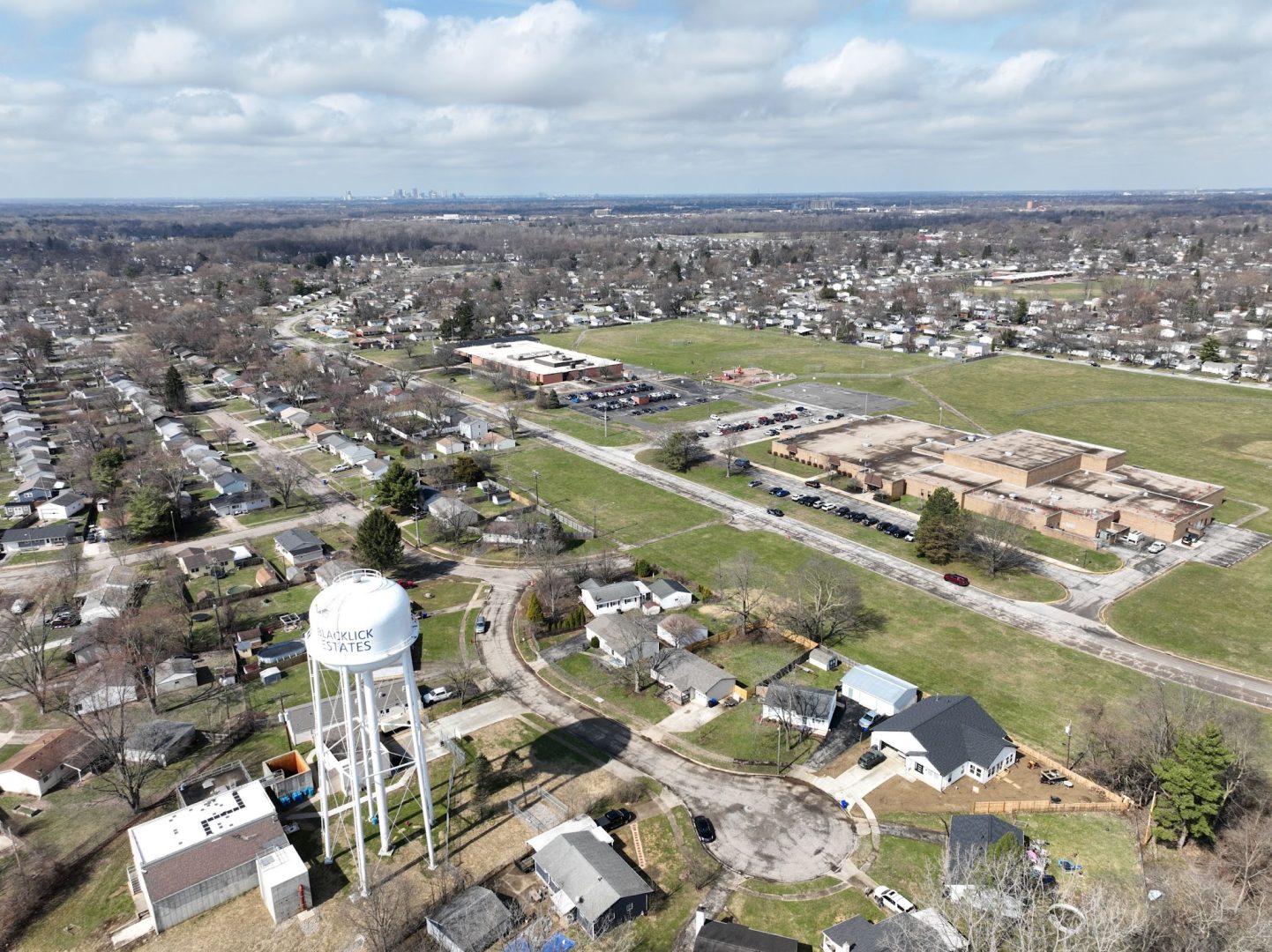 Aerial view of a suburban neighborhood with a white water tower labeled SAVAGE 1947. Nearby are houses, a large school building, parking lots, open green spaces, and distant city skyline under a partly cloudy sky.