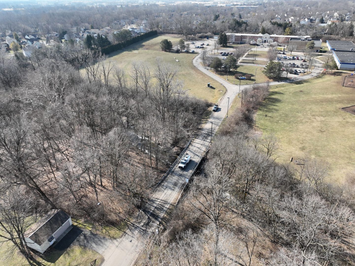 Aerial view of a car crossing a bridge over a wooded area, with open fields, a few buildings, and a parking lot in the background on a clear day.