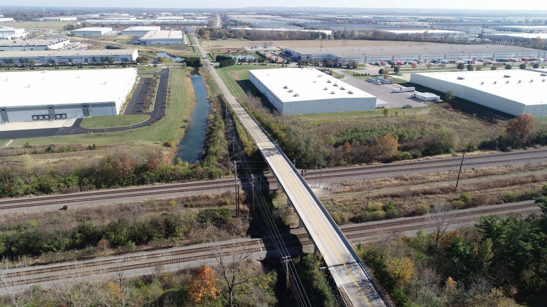 Aerial view of an industrial area with large white warehouse buildings, a canal, and railway tracks crossed by a road bridge, surrounded by grassy and wooded areas under a clear sky.