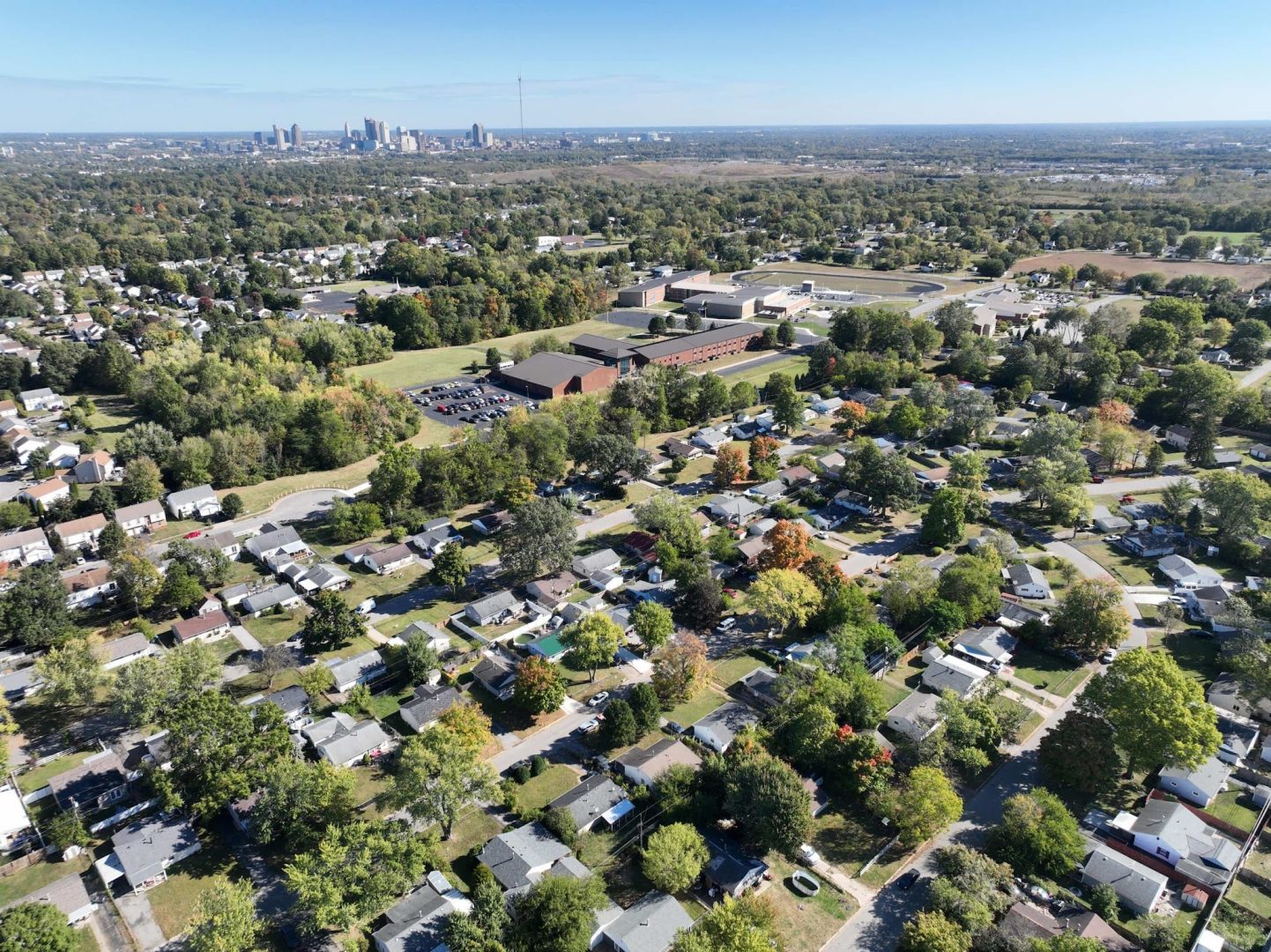 Aerial view of a suburban neighborhood with tree-lined streets and single-family homes, a large school complex in the center, and a distant city skyline under a clear blue sky.