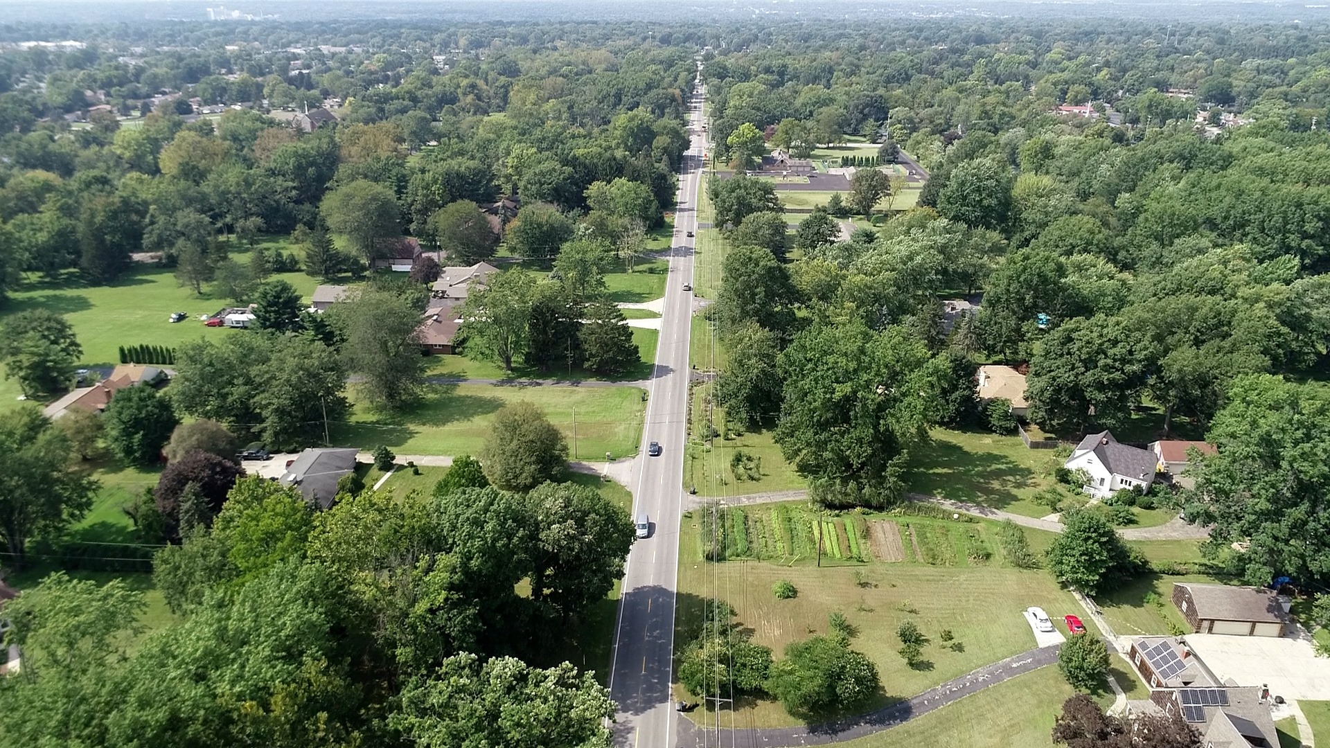 Aerial view of a straight road running through a suburban neighborhood with houses, dense trees, and green yards on both sides, stretching into the distance under a hazy sky. Aerial view of a straight road running through a suburban neighborhood with houses, dense trees, and green yards on both sides, stretching into the distance under a hazy sky.