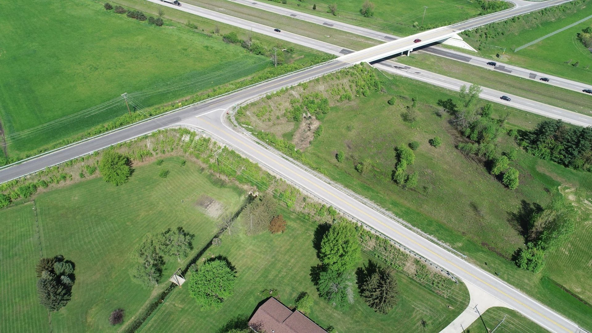 Aerial view of a rural road intersection with an overpass, surrounded by green fields, trees, and a single house in the bottom corner. Cars are visible on the highway and the landscape appears well-maintained.