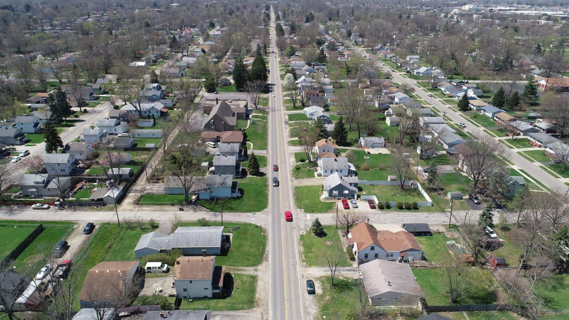 Aerial view of a suburban neighborhood with a straight road running down the center, lined with houses, green lawns, trees, and a few cars driving and parked along the street. Aerial view of a suburban neighborhood with a straight road running down the center, lined with houses, green lawns, trees, and a few cars driving and parked along the street.