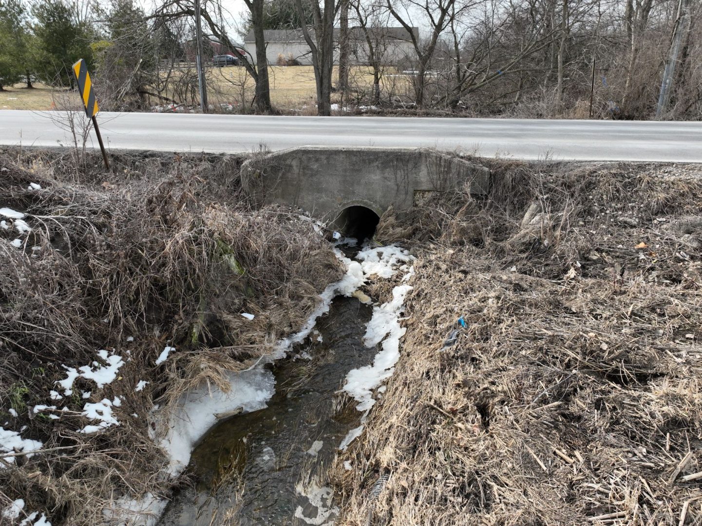 A small creek flows under a concrete culvert beneath a rural road. Sparse snow patches remain on the ground and dry grass, with leafless trees and a building in the background.