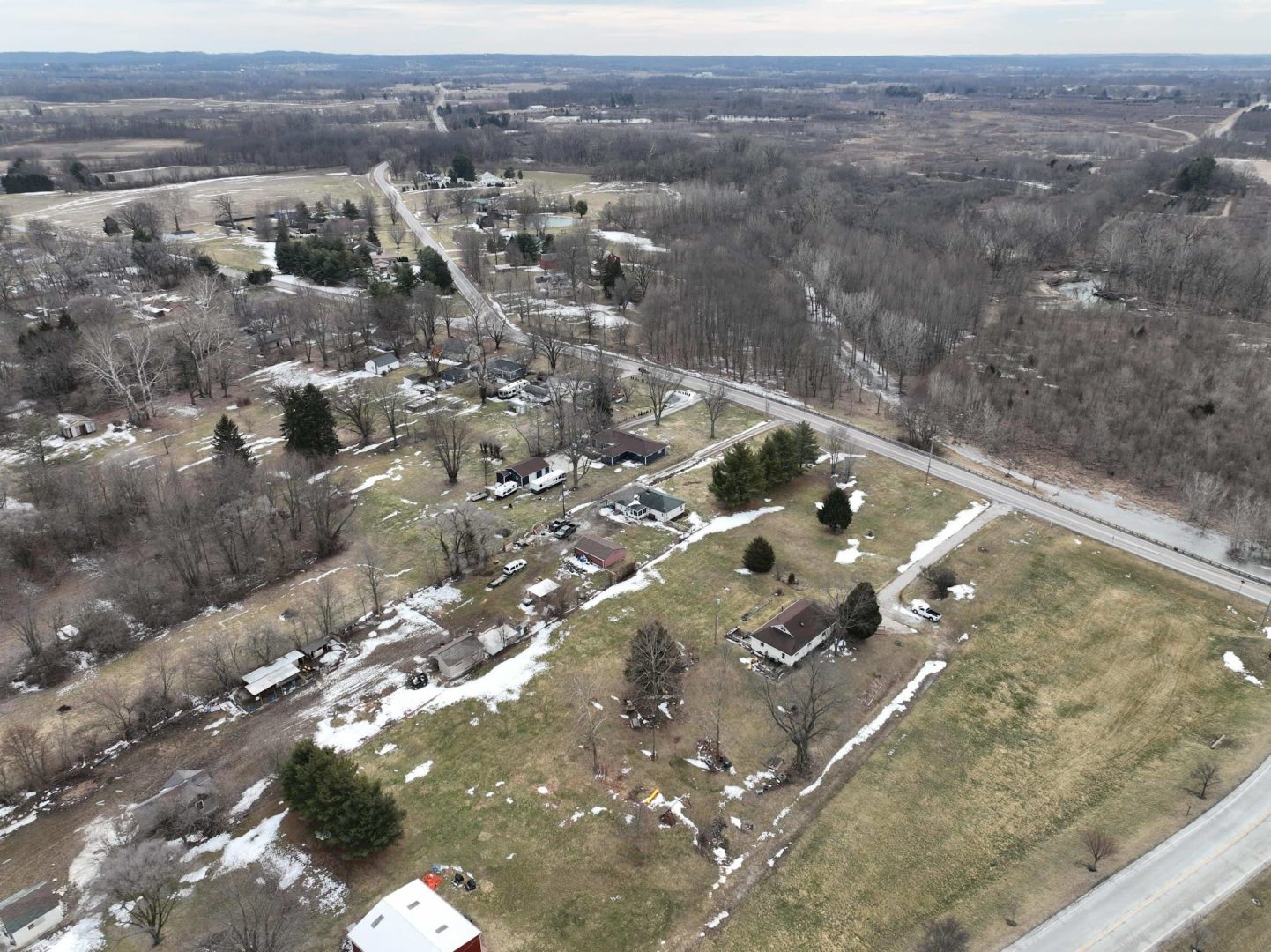 Aerial view of a rural area with scattered houses, patches of melting snow, trees without leaves, and winding roads. The landscape is mostly brown and green, with fields and wooded areas under a cloudy sky. Aerial view of a rural area with scattered houses, patches of melting snow, trees without leaves, and winding roads. The landscape is mostly brown and green, with fields and wooded areas under a cloudy sky.