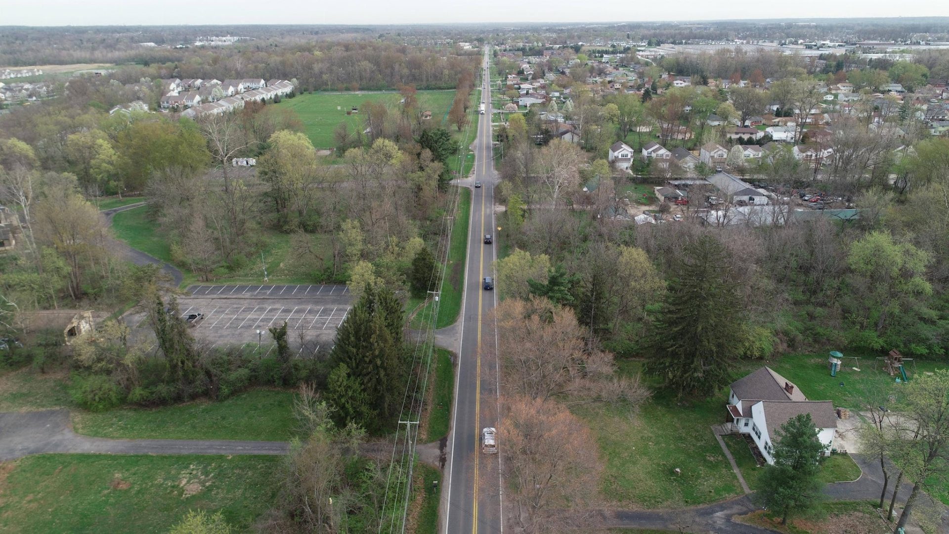Aerial view of a long, straight road running through a suburban area with trees, houses, a parking lot, and grassy fields on either side under an overcast sky. Aerial view of a long, straight road running through a suburban area with trees, houses, a parking lot, and grassy fields on either side under an overcast sky.