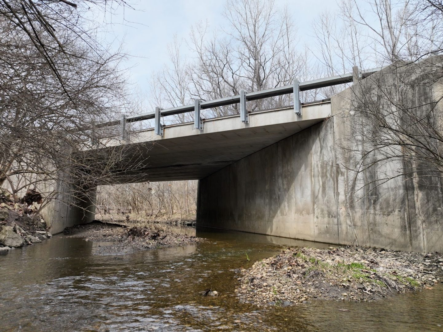 A concrete bridge spans over a shallow creek with bare trees on both sides. The sky is overcast, and there are rocks and patches of grass along the water’s edge.