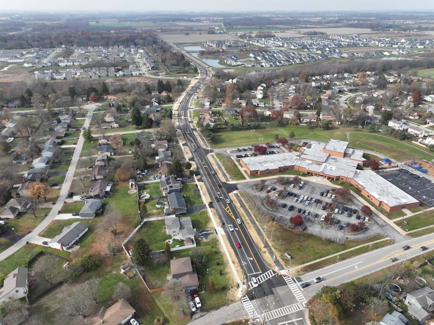 Aerial view of a suburban neighborhood with houses, a school with a parking lot, streets with vehicles, and open fields in the background. Trees with autumn foliage are scattered throughout the area. Aerial view of a suburban neighborhood with houses, a school with a parking lot, streets with vehicles, and open fields in the background. Trees with autumn foliage are scattered throughout the area.
