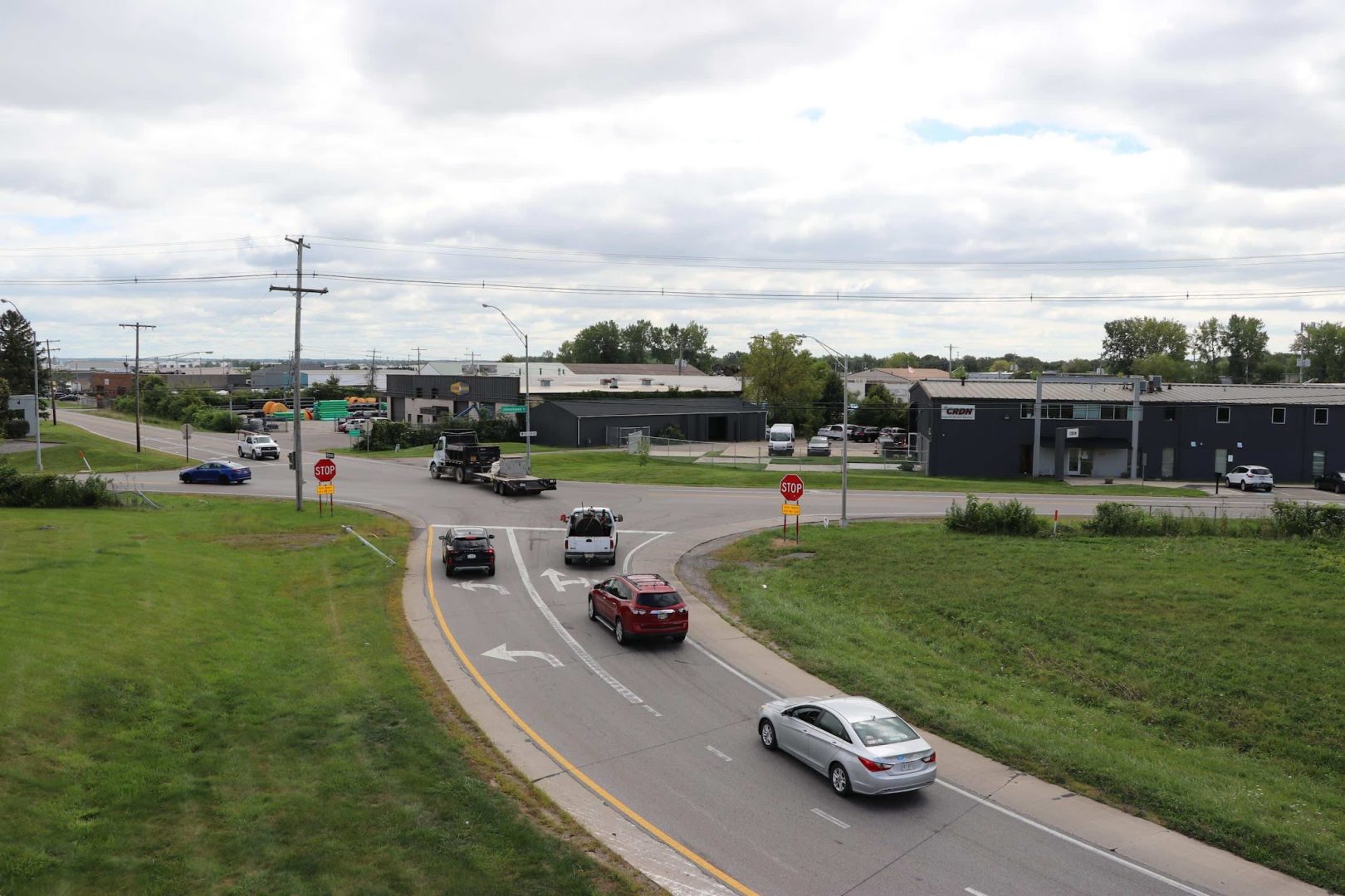 Cars drive on a curving road toward an intersection with stop signs. Buildings and grassy areas are visible under a cloudy sky. Some vehicles are waiting at the intersection while others are merging onto the road. Cars drive on a curving road toward an intersection with stop signs. Buildings and grassy areas are visible under a cloudy sky. Some vehicles are waiting at the intersection while others are merging onto the road.