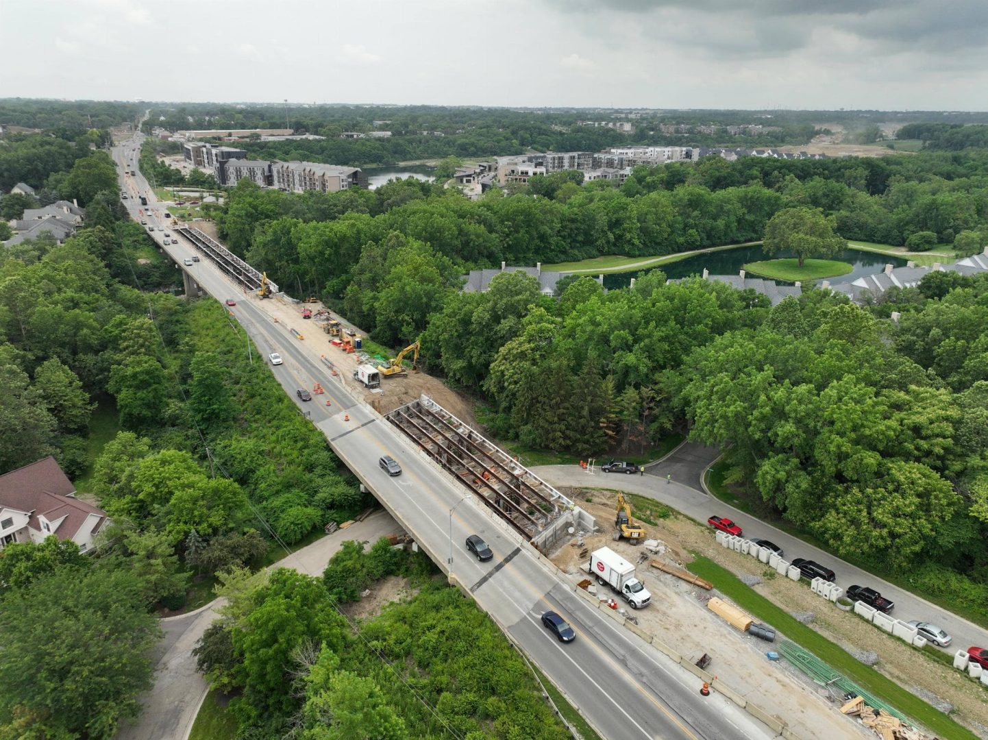 Aerial view of a road bridge under construction, with vehicles passing on partially completed lanes, surrounded by green trees, residential buildings, and cloudy skies overhead. Construction equipment and workers are visible near the site.