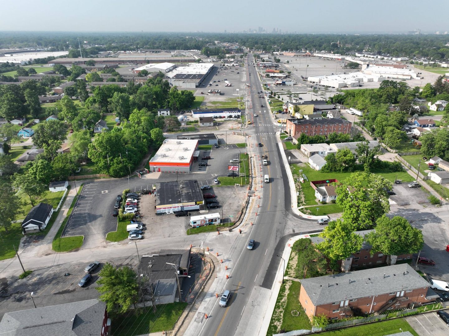 Aerial view of a busy urban street lined with businesses, stores, parking lots, and residential buildings, with trees scattered throughout and cars driving along the road under a hazy sky.