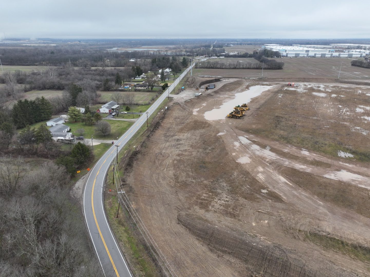 A rural road curves past a residential area with houses and trees, next to a large muddy construction site with heavy equipment and puddles, under an overcast sky. A rural road curves past a residential area with houses and trees, next to a large muddy construction site with heavy equipment and puddles, under an overcast sky.