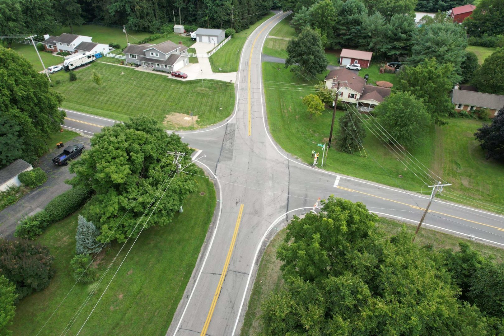 Aerial view of a rural intersection with three roads meeting, surrounded by green lawns, trees, several houses, and driveways. Power lines cross the roads and properties. The area appears calm and well-kept. Aerial view of a rural intersection with three roads meeting, surrounded by green lawns, trees, several houses, and driveways. Power lines cross the roads and properties. The area appears calm and well-kept.
