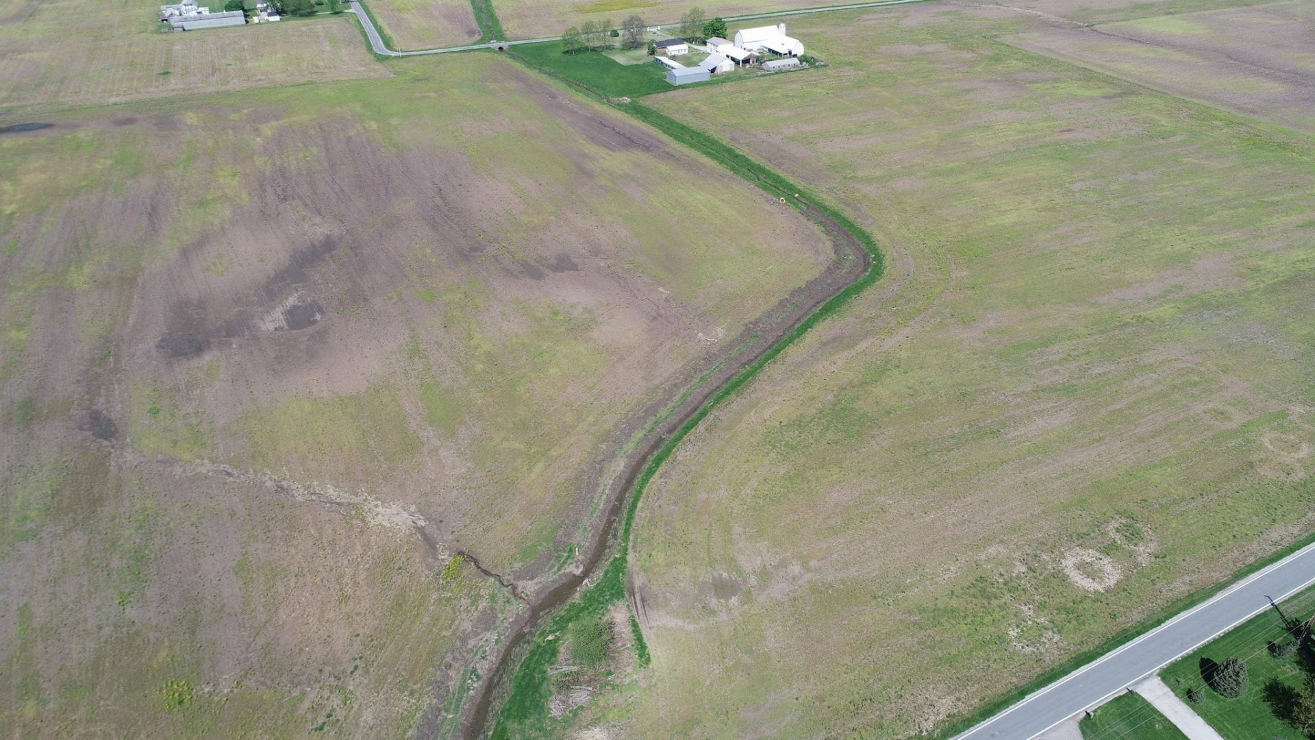 Aerial view of a large, mostly empty green field with a winding dirt path or stream running through it, a few buildings near the top, and a road along the bottom right edge.