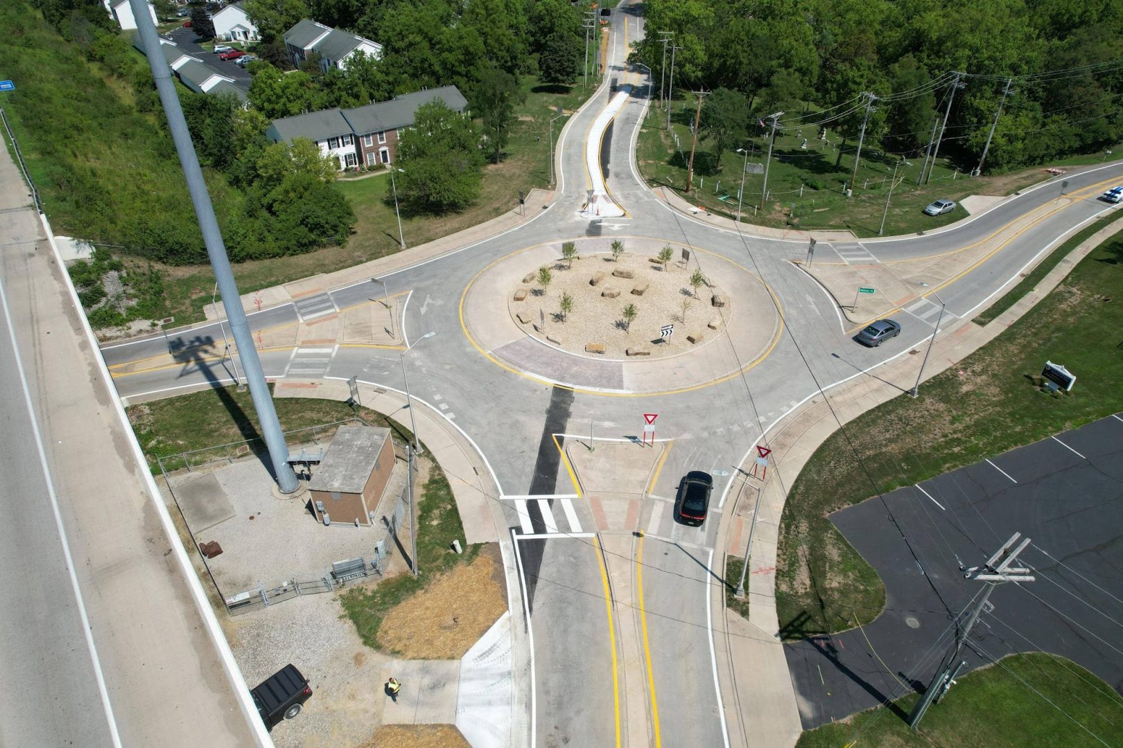 Aerial view of a roundabout intersection with several cars navigating, crosswalks, landscaped center, nearby buildings, trees, and a parking lot. Multiple roads connect to the roundabout.