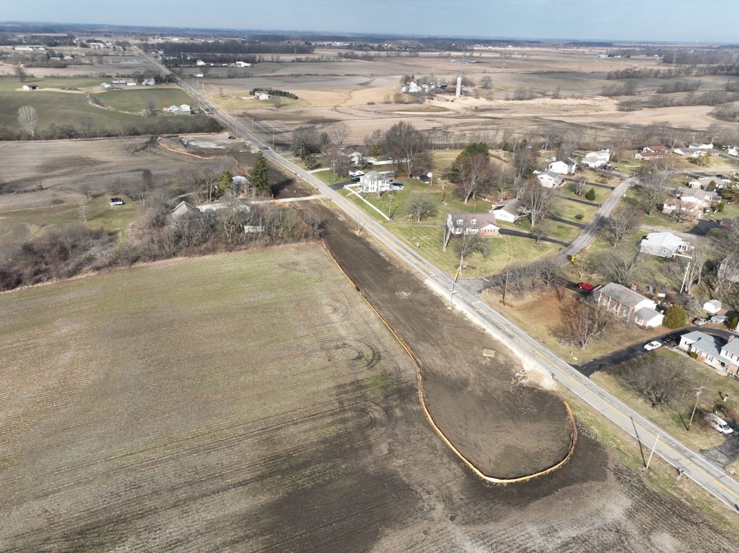 Aerial view of a rural landscape showing a road running through fields and a small neighborhood, with construction work and soil erosion barriers visible alongside the road. Sparse trees and farm buildings are scattered in the distance. Aerial view of a rural landscape showing a road running through fields and a small neighborhood, with construction work and soil erosion barriers visible alongside the road. Sparse trees and farm buildings are scattered in the distance.