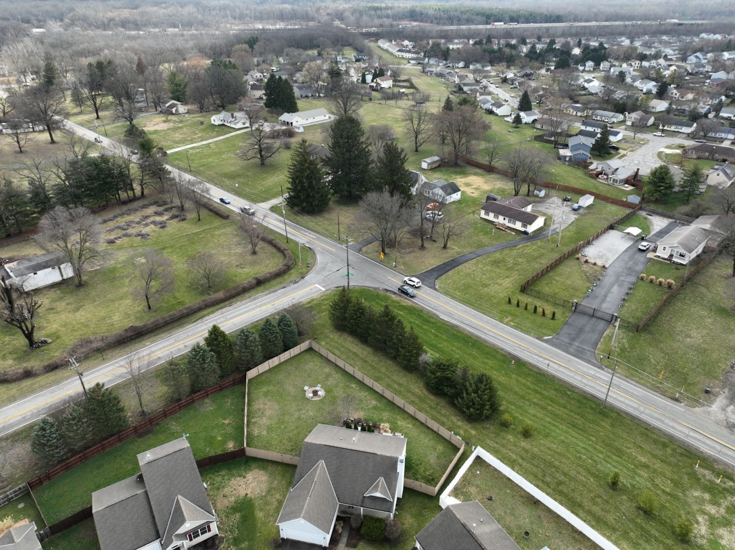 Aerial view of a suburban neighborhood with intersecting roads, houses with yards, trees, and open grassy areas. Some driveways and fences are visible, and the overall scene looks calm and spacious.
