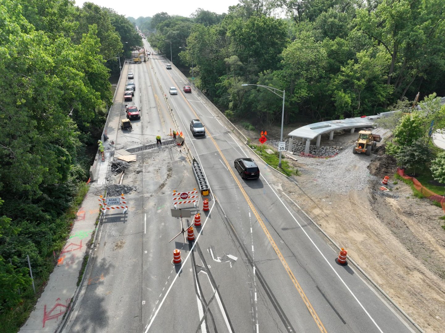 Aerial view of a road construction site with traffic cones, barriers, construction workers, and vehicles. One lane is closed, and construction equipment is visible near a partially built structure beside the road. Trees line both sides.