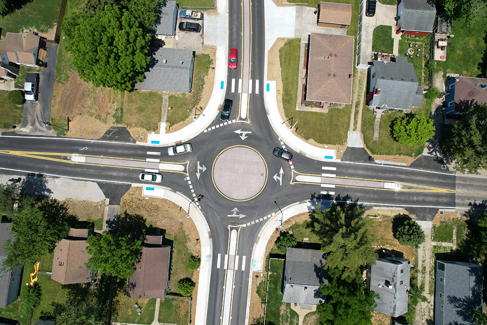 Aerial view of a roundabout at a residential intersection with cars navigating the circle, surrounded by houses, trees, crosswalks, and sidewalks on all sides.