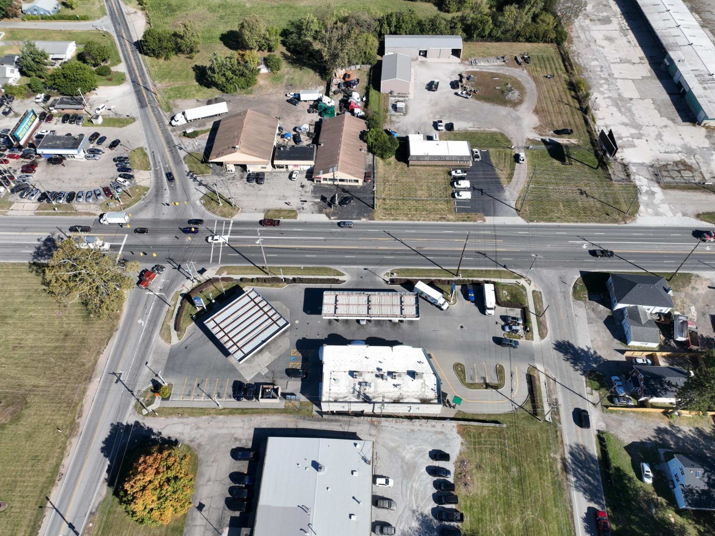 Aerial view of a commercial area with multiple buildings, parking lots, cars, and a main road running horizontally through the center, surrounded by grassy areas and some trees.