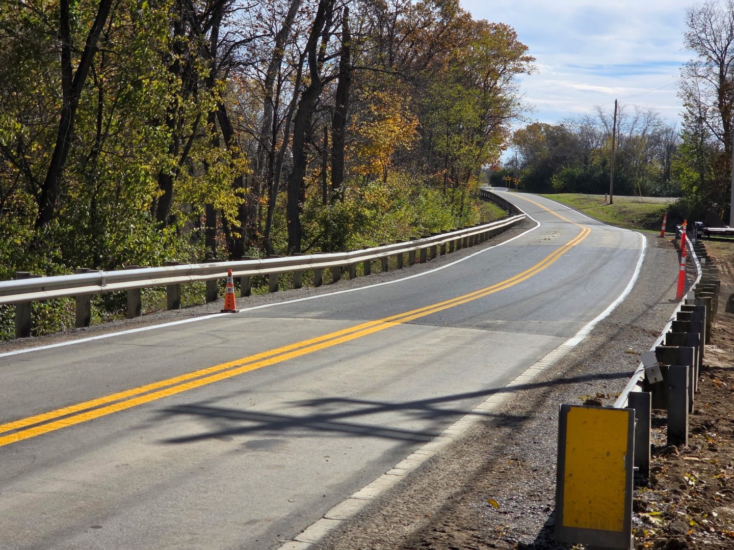 A two-lane road with yellow lines curves through a wooded area. Guardrails run along both sides, and an orange traffic cone marks the shoulder. Trees with autumn leaves line the road under a clear sky.
