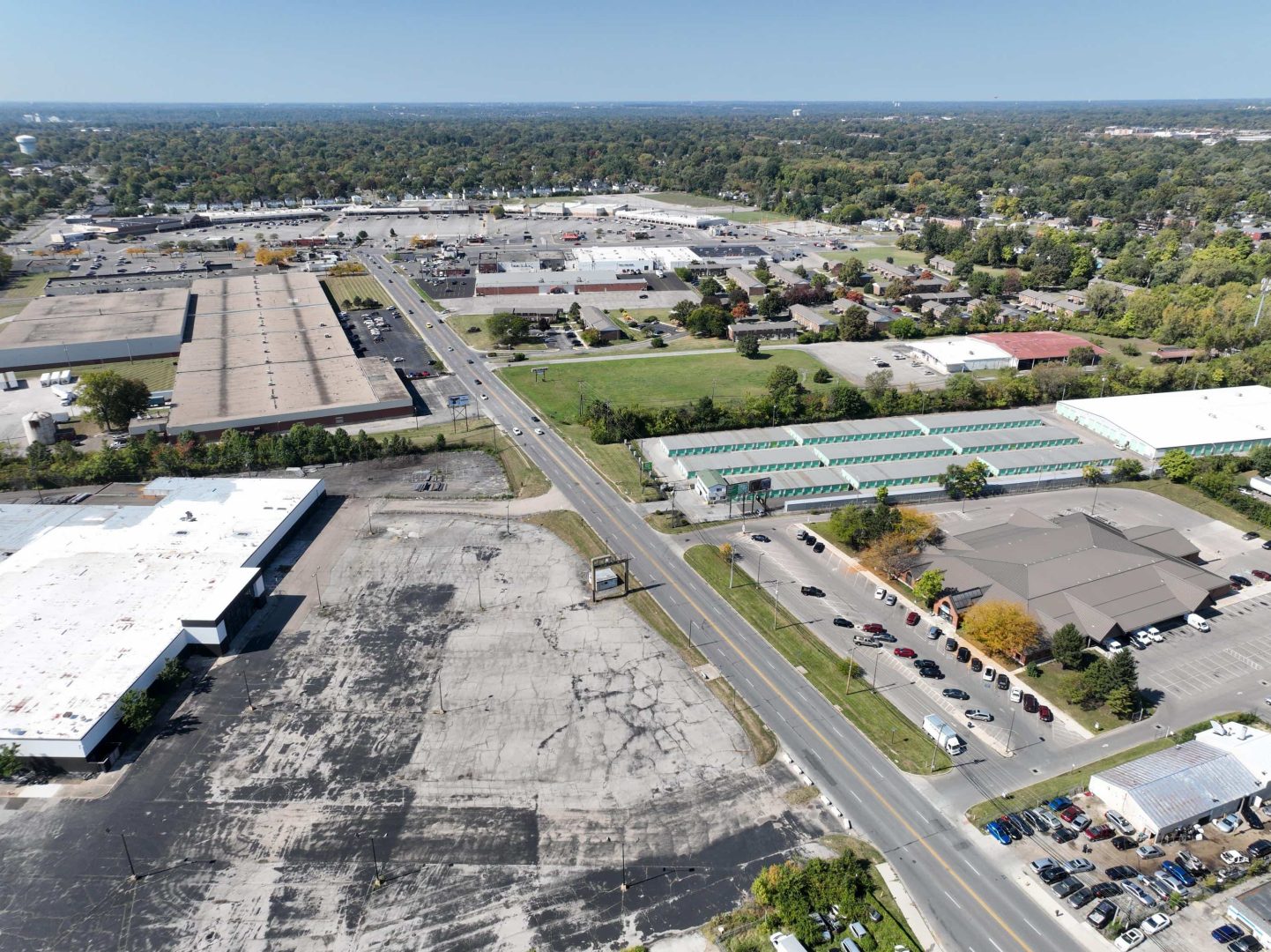 Aerial view of a commercial area with warehouses, parking lots, roads, and scattered trees. Several cars are parked, and a few drive along the main road running through the center of the image.