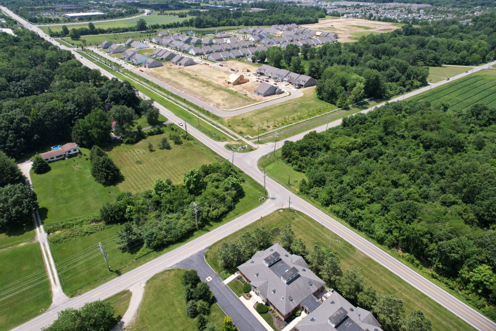 Aerial view of a suburban area with a large housing development under construction, surrounded by green trees and roads intersecting in the foreground, with homes and open fields nearby.
