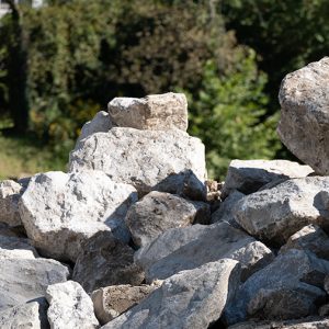 A pile of large, irregularly-shaped gray stones and rocks sits outdoors, with green trees and foliage visible in the blurred background on a sunny day.