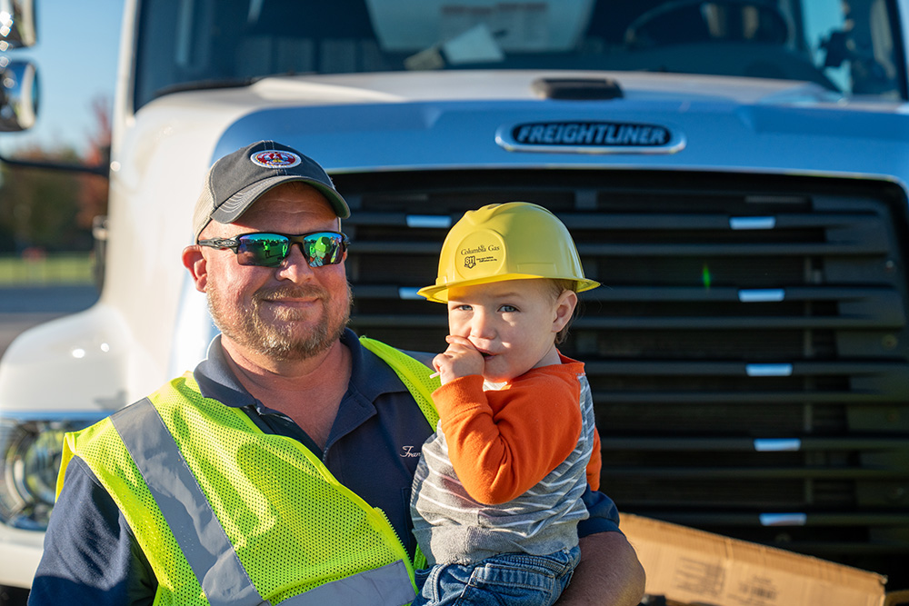 A man in a neon safety vest and sunglasses holds a smiling toddler wearing a yellow toy hard hat in front of a Freightliner truck.