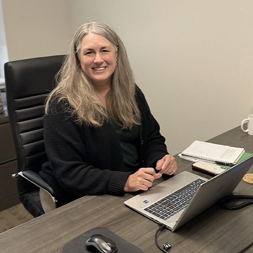 A woman with long gray hair sits at a desk, smiling at the camera. She is wearing a black cardigan and has a laptop, notebook, mug, and smartphone in front of her in an office setting.