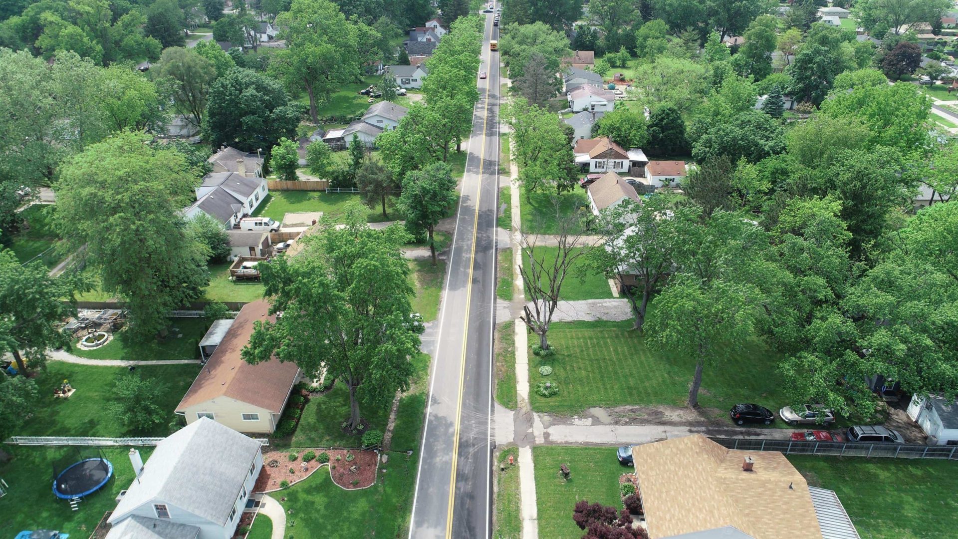 Aerial view of a residential neighborhood with tree-lined streets, houses with yards, parked cars, and a central road running vertically through the image. Lush green trees and lawns surround the homes. Aerial view of a residential neighborhood with tree-lined streets, houses with yards, parked cars, and a central road running vertically through the image. Lush green trees and lawns surround the homes.