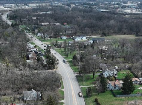 Aerial view of a two-lane road passing through a suburban area with houses, trees, and sparse traffic on a cloudy day. The landscape is mostly bare, with leafless trees and open green spaces.