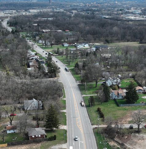 Aerial view of a two-lane road passing through a suburban area with houses, trees, and sparse traffic on a cloudy day. The landscape is mostly bare, with leafless trees and open green spaces.