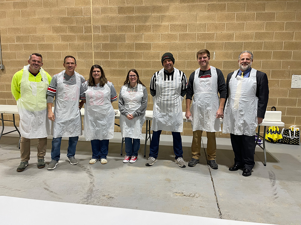 Seven people stand side by side indoors, wearing white aprons over their clothes, in front of a brick wall and several folding tables. They are posing and smiling for the photo in a casual setting.