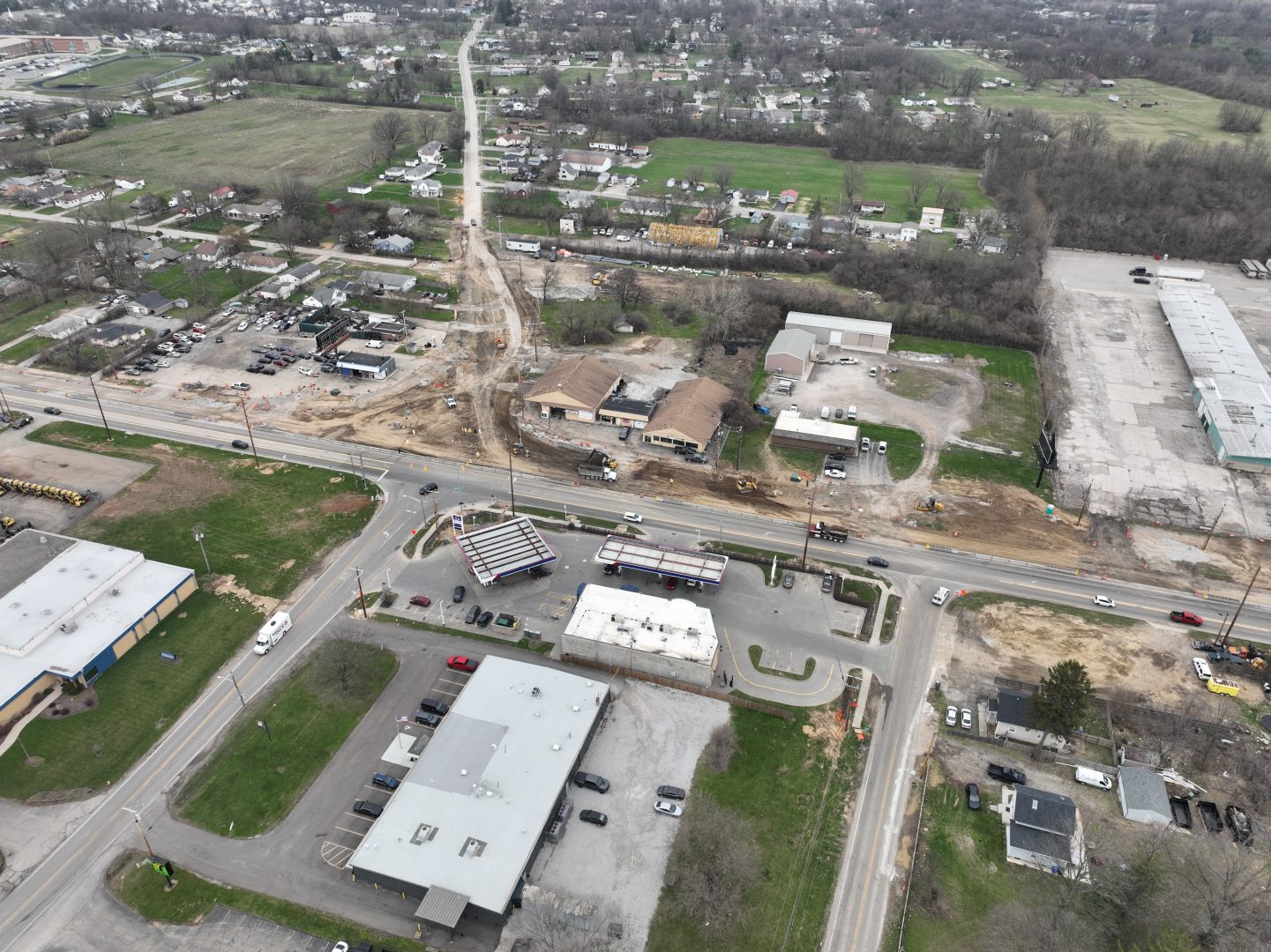 Aerial view of a suburban area with buildings, roads, and parked cars. A construction site with cleared land and equipment is visible near the center, and residential houses and fields surround the area. Trees and greenery are in the background. Aerial view of a suburban area with buildings, roads, and parked cars. A construction site with cleared land and equipment is visible near the center, and residential houses and fields surround the area. Trees and greenery are in the background.