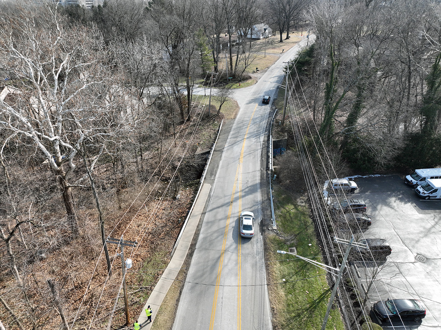Aerial view of a two-lane road bordered by leafless trees, a sidewalk, and utility poles. Two cars drive along the road, and a few parked vehicles are visible in a nearby parking lot. Aerial view of a two-lane road bordered by leafless trees, a sidewalk, and utility poles. Two cars drive along the road, and a few parked vehicles are visible in a nearby parking lot.