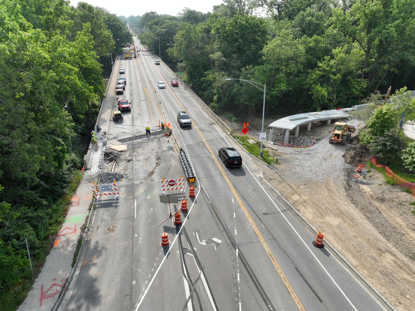 Aerial view of a road construction site with traffic cones, barriers, and workers. Vehicles drive through one open lane; heavy machinery operates near a bus stop shelter on the right, surrounded by trees and greenery. Aerial view of a road construction site with traffic cones, barriers, and workers. Vehicles drive through one open lane; heavy machinery operates near a bus stop shelter on the right, surrounded by trees and greenery.
